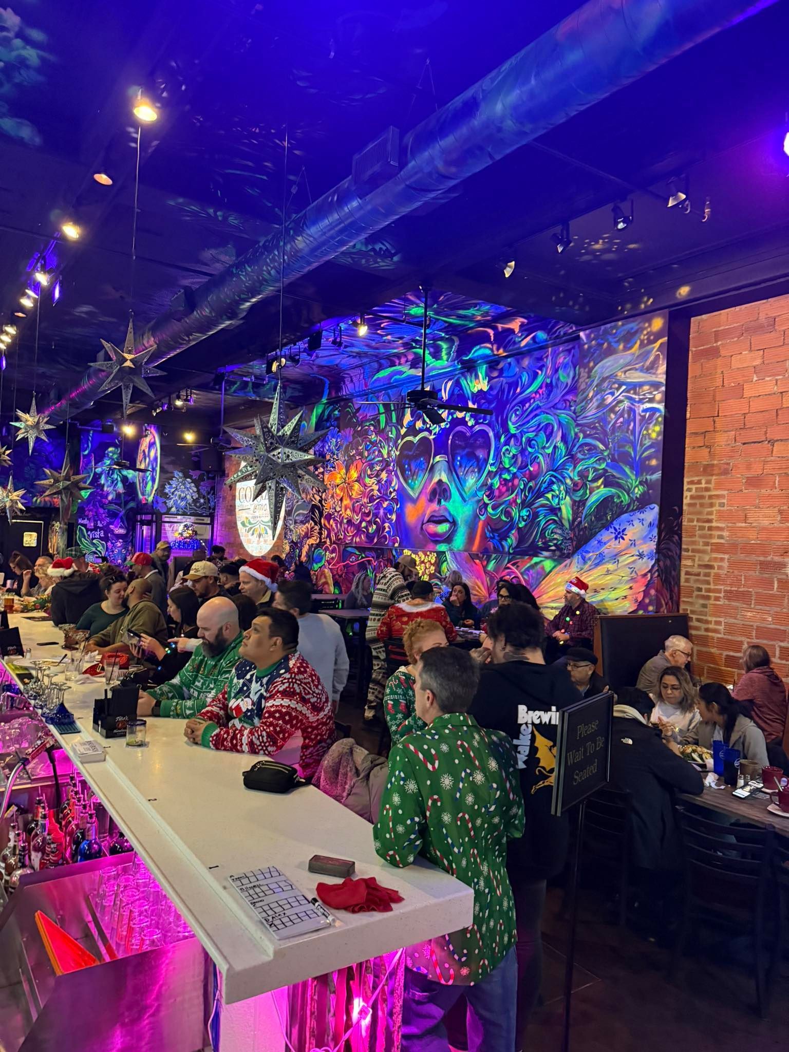 People at a bar with colorful mural, Christmas decorations, and festive sweaters.