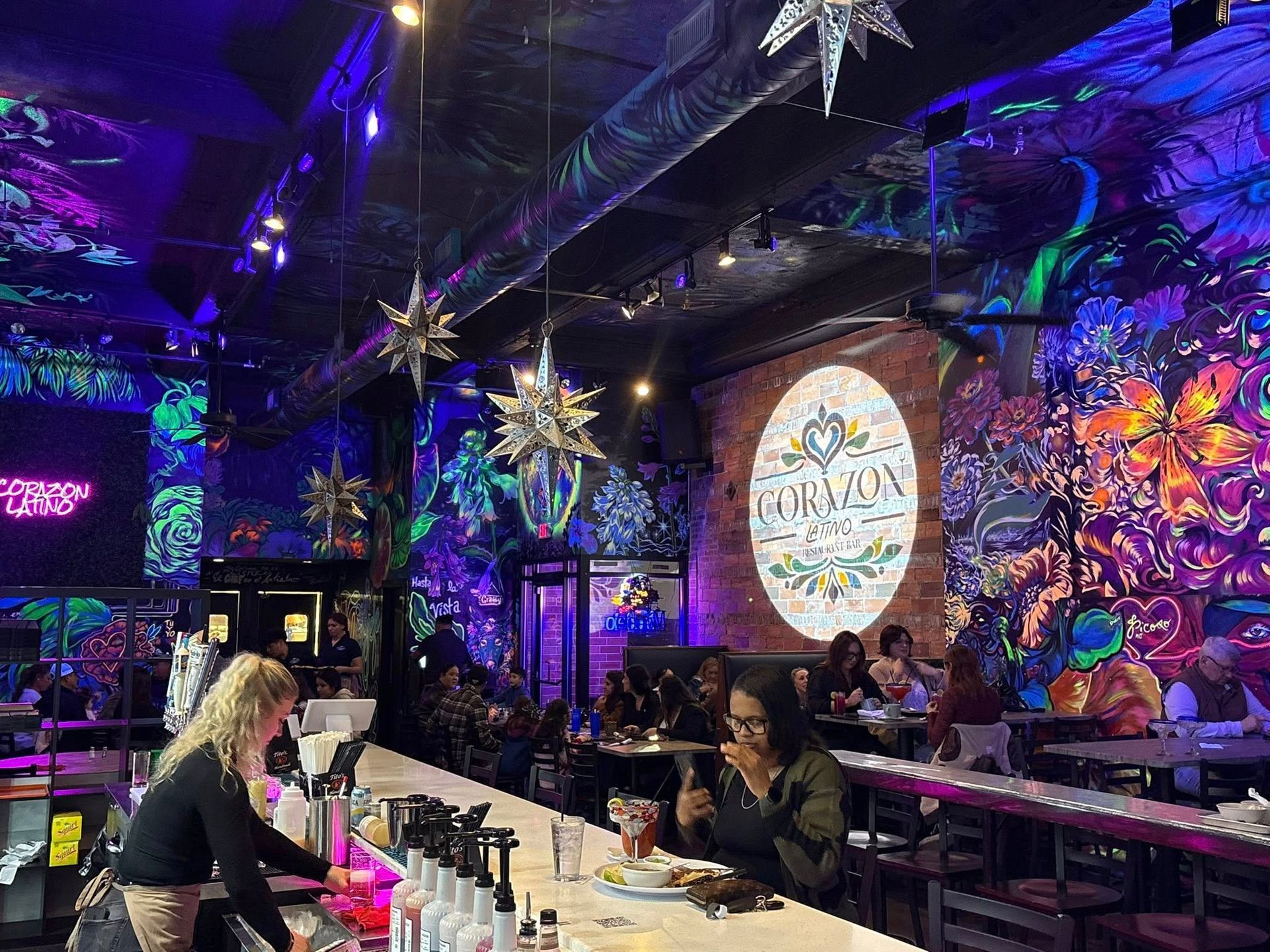 Bar interior with neon art on walls, a bar, patrons seated at tables, and a bartender.
