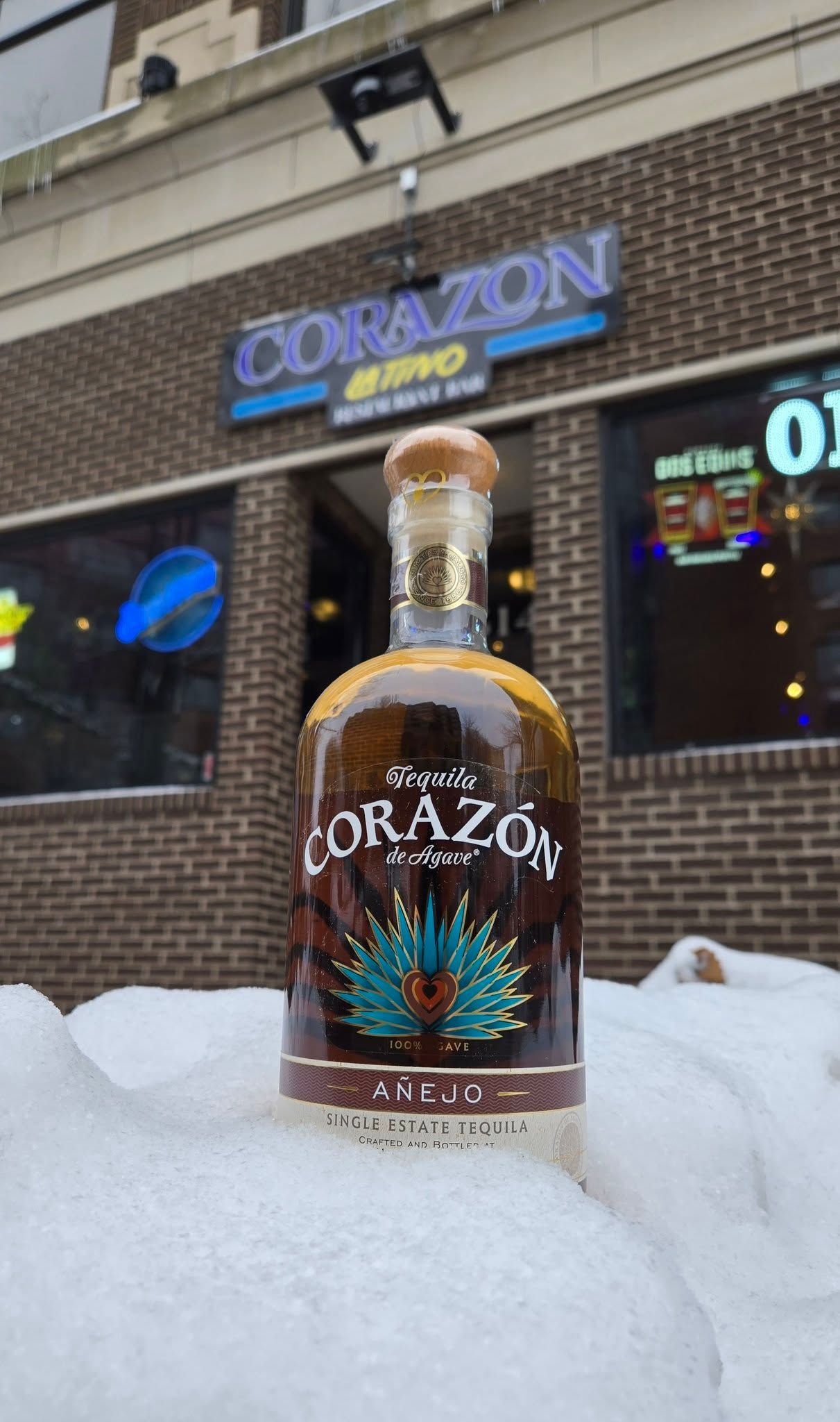 Bottle of Corazón tequila on snow in front of Corazón Bistro, a brick building with a blue sign.