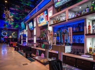 Bar interior with neon blue and green lights, multiple TVs, liquor bottles on shelves, and a long bar.