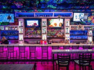 A vibrant bar with pink neon lights. Television screens and liquor bottles are displayed behind the bar.