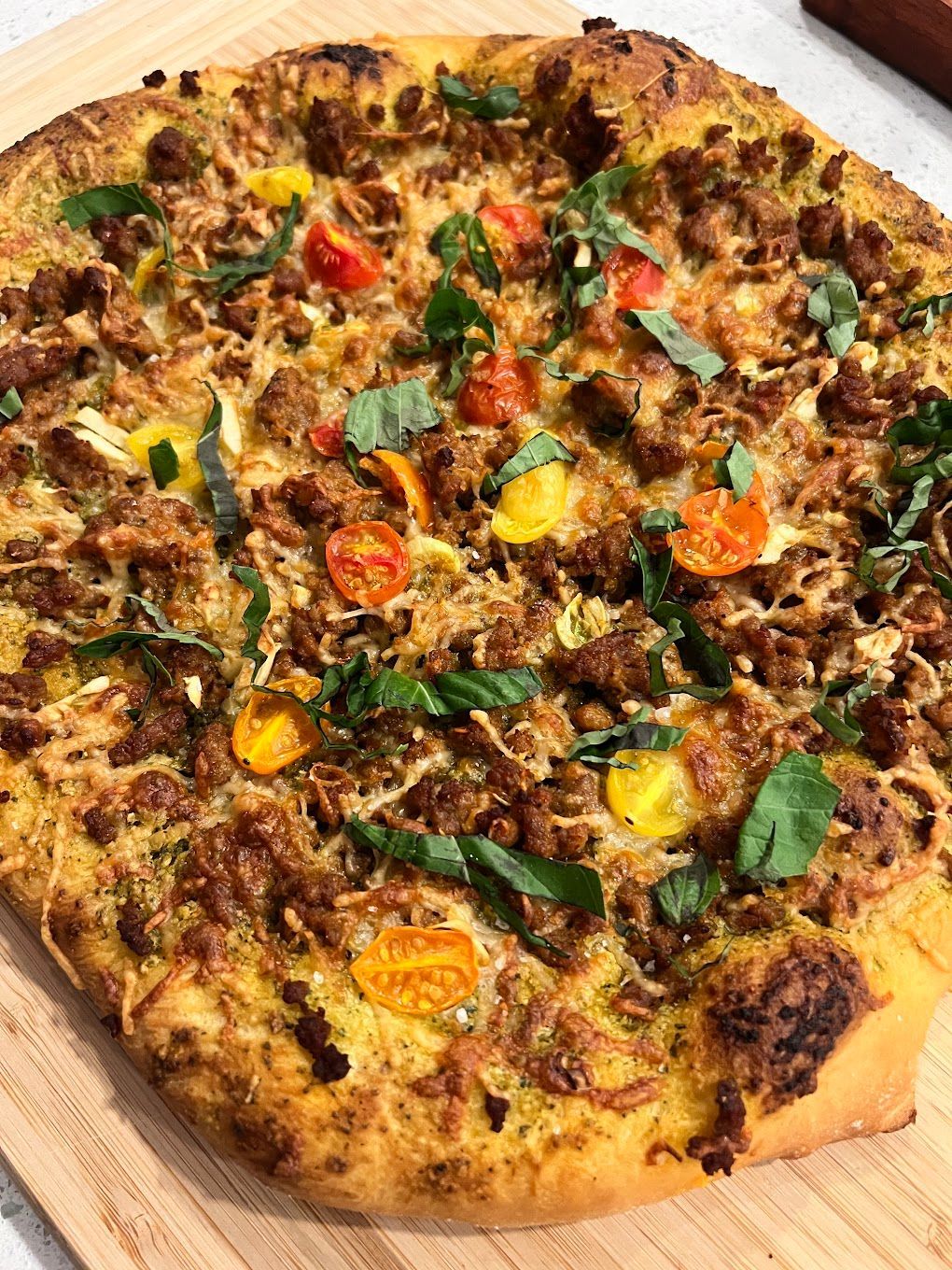 A pizza with meat , tomatoes , basil and cheese is sitting on a wooden cutting board.