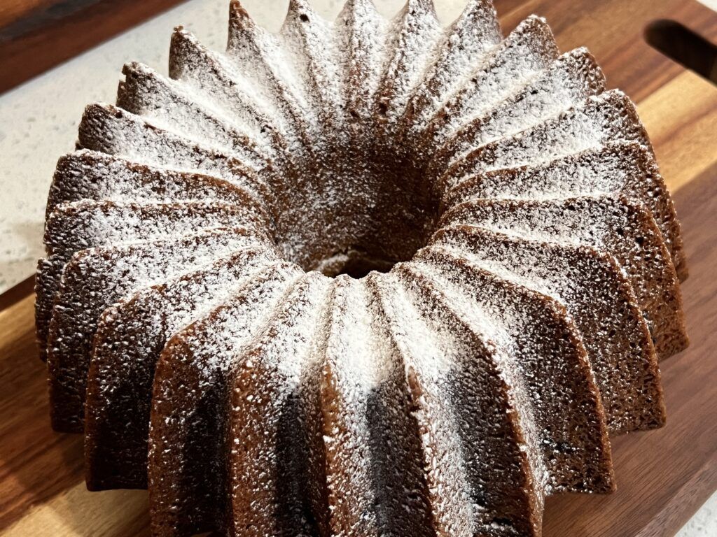 A bundt cake covered in powdered sugar is sitting on a wooden cutting board.