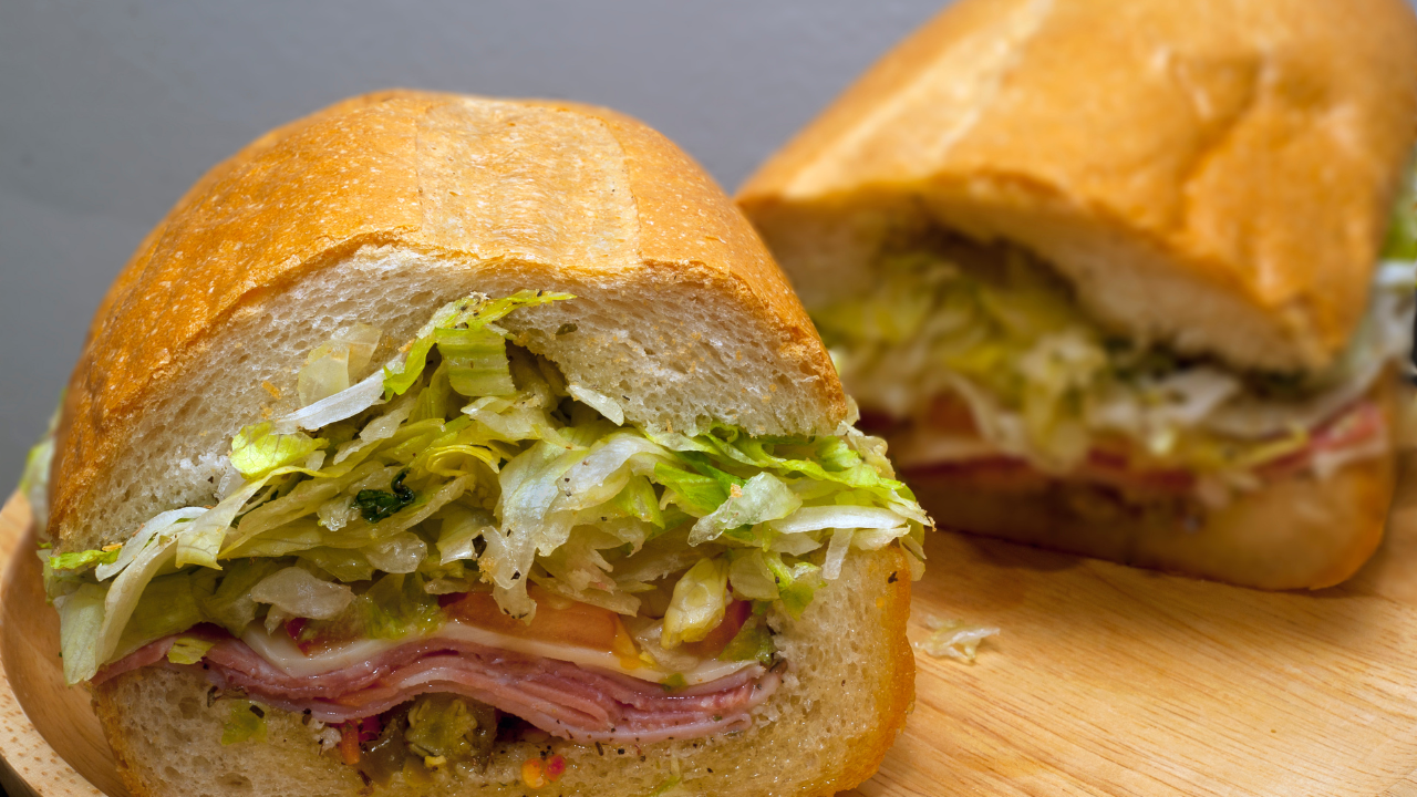 Two sub sandwiches are cut in half on a wooden cutting board.