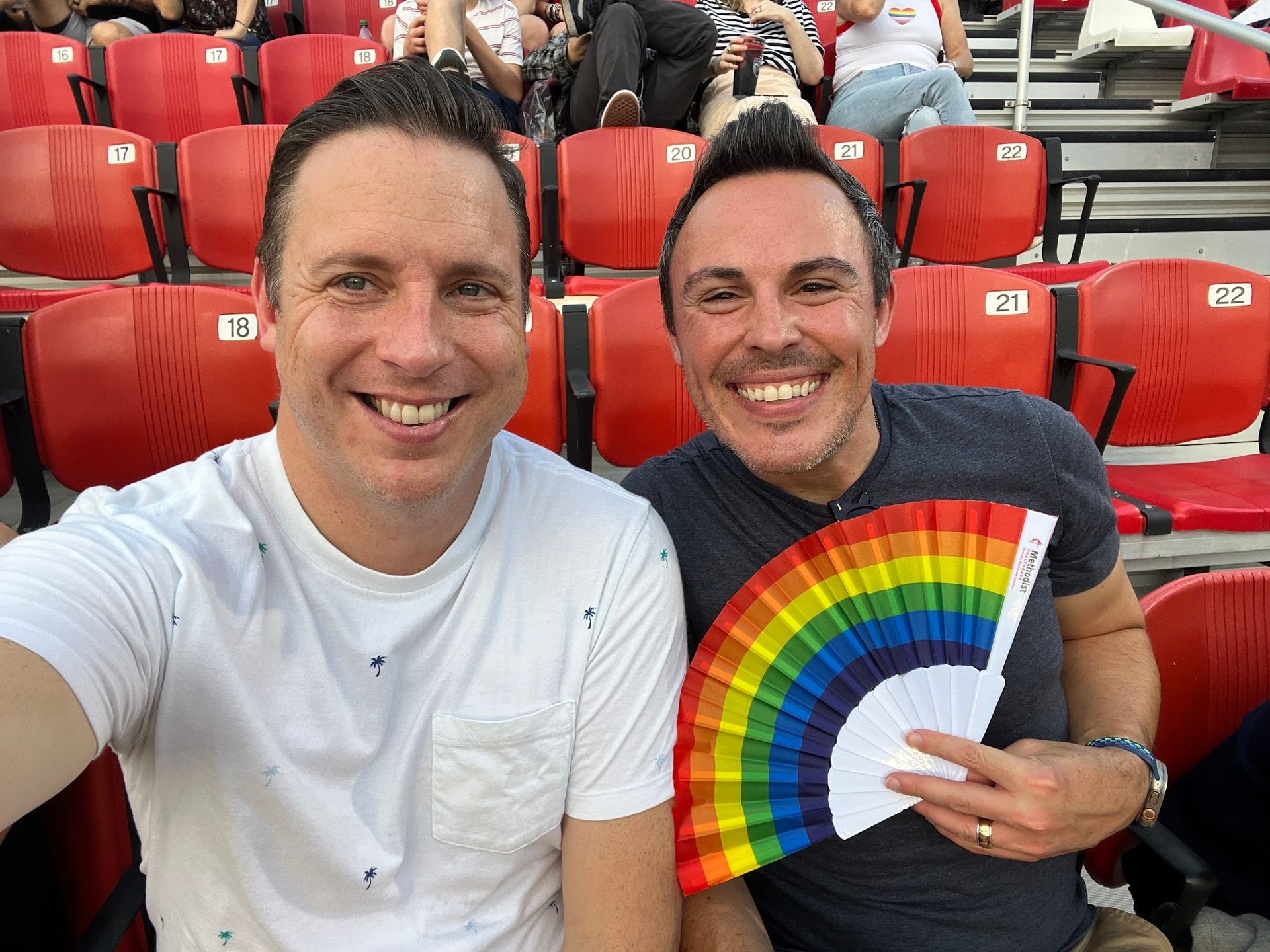 Two men are sitting in a stadium holding a rainbow fan.