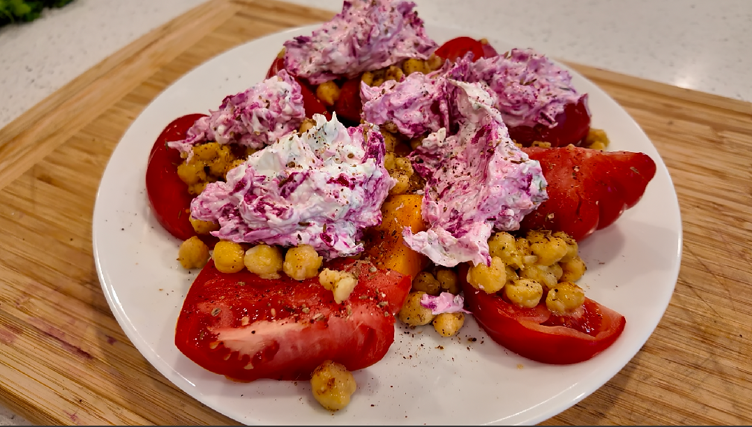 A white plate topped with tomatoes , chickpeas and purple sauce on a wooden cutting board.