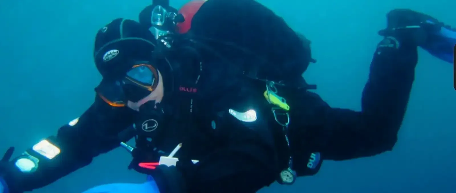 Scuba diver in black gear underwater, holding a camera and flashing a hand signal