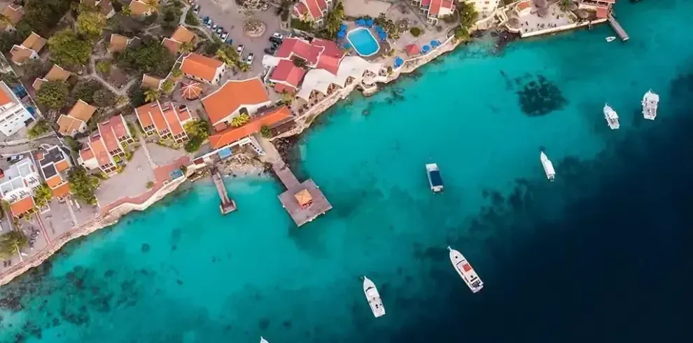 Aerial view of a seaside resort with turquoise water, boats, and red-roofed buildings along the coast