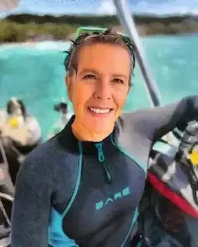 Smiling person in a black wetsuit on a boat, with turquoise water and shoreline in the background