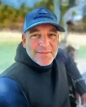 Man wearing a blue cap and black jacket smiling outdoors near the beach