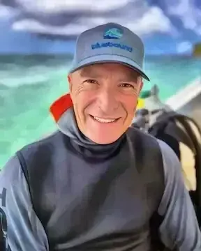 Smiling person in a gray hoodie and blue cap sitting by turquoise water and cloudy sky