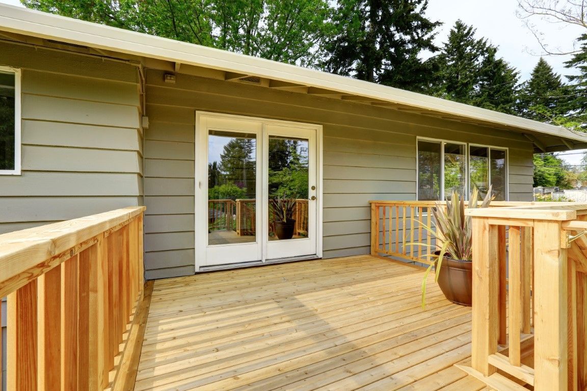 Wooden deck leading to beige house with white French doors and railing, surrounded by trees