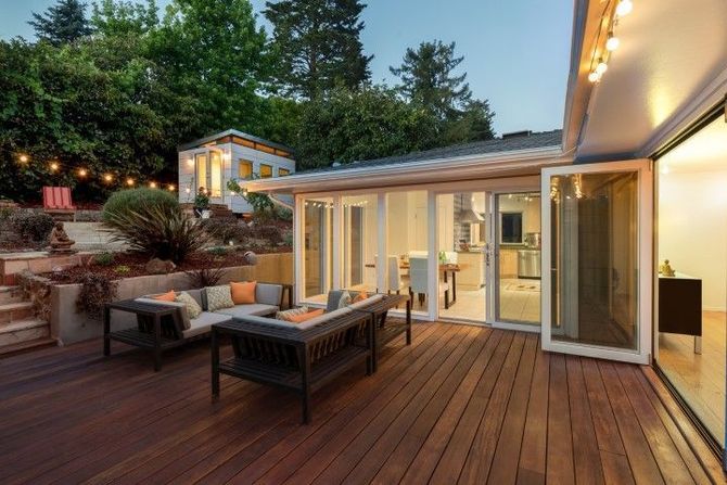 Backyard deck with outdoor seating beside a lit glass-walled house at dusk