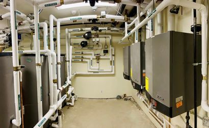 Water softener and filtration tanks in a basement near exposed pipes and a utility sink.