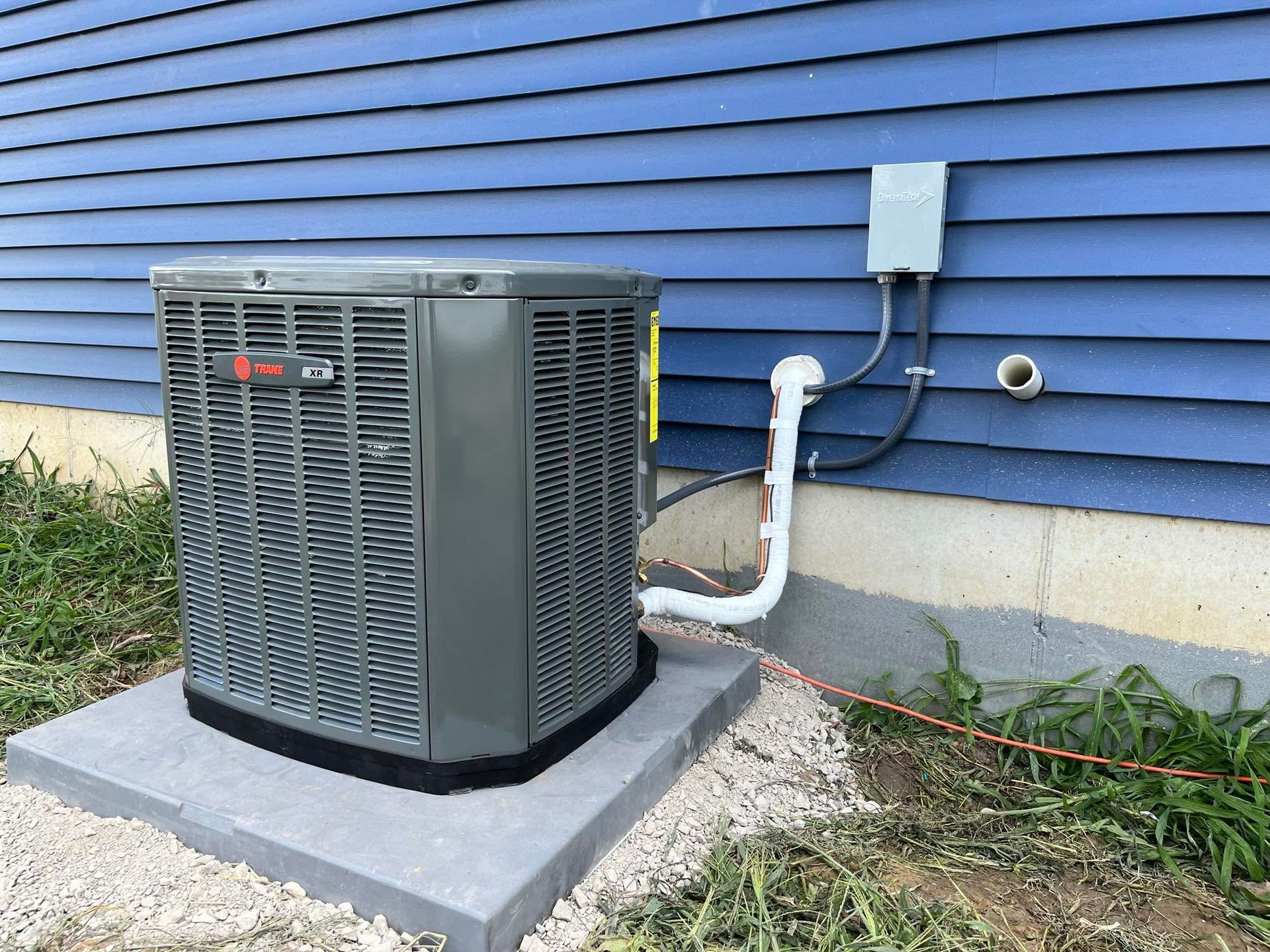 Outdoor air conditioner unit beside a blue house, with white piping and concrete pad.