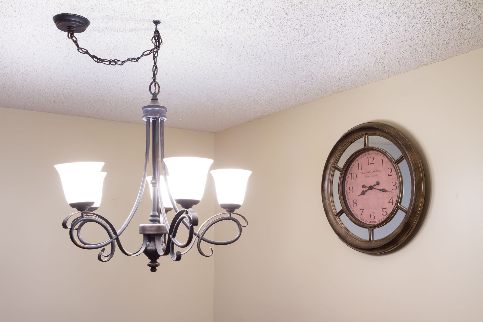 Ceiling chandelier with lit frosted shades beside a round wall clock in a beige room