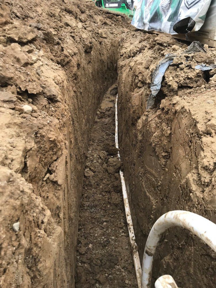 Deep narrow trench in muddy ground with a pipe running along the bottom, workers visible at the top edge