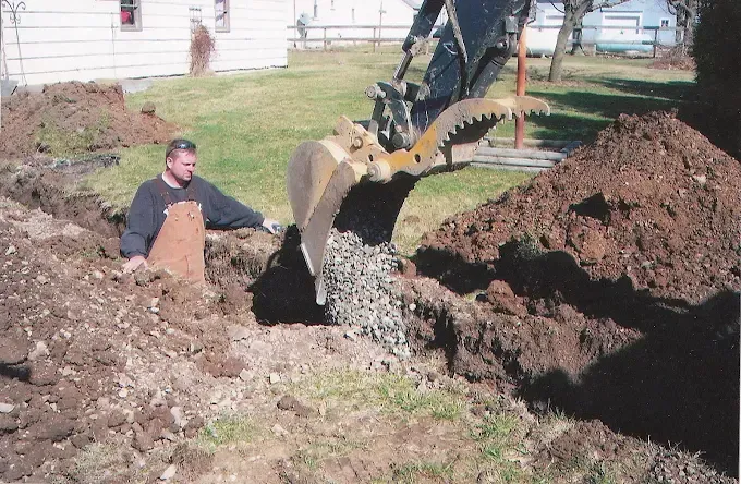 Man in a dirt trench beside an excavator digging near a house, with large soil piles around him