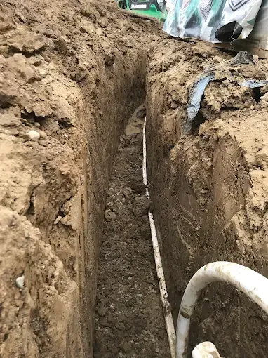 Deep trench in dirt with a white pipe running along the bottom at a construction site