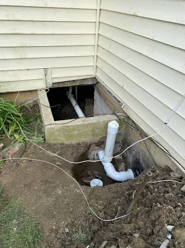 Open crawlspace vent and exposed white PVC pipes beside a house foundation.