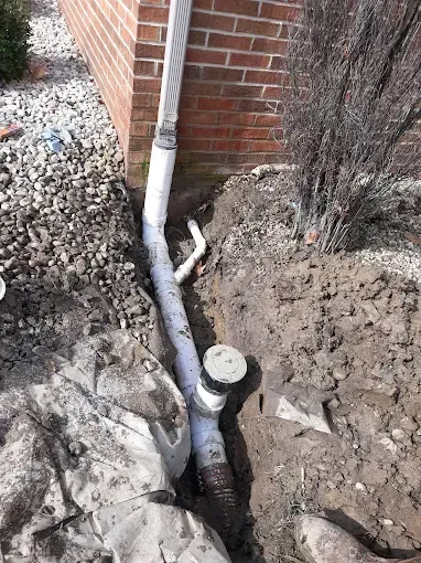 Exposed PVC pipes and a capped drain line beside a brick house in a muddy excavation.