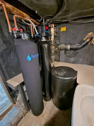 Water softener and filtration tanks in a basement near exposed pipes and a utility sink.
