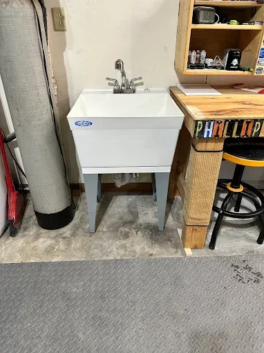 White utility sink beside a wooden workbench in a garage-like room with a gray floor mat.