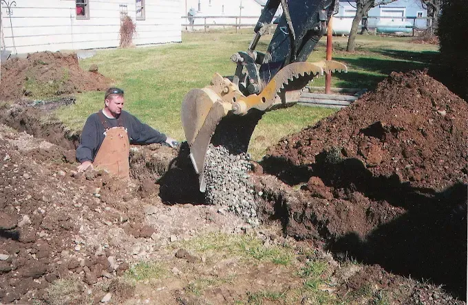 Man sitting beside an excavator digging a trench in a backyard with dirt piles