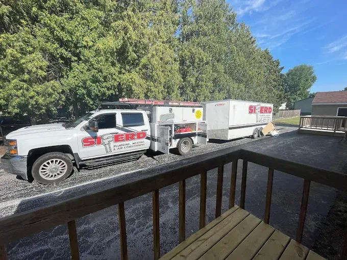 White utility truck with red lettering and trailer parked on a gravel lot by trees, seen from a porch.