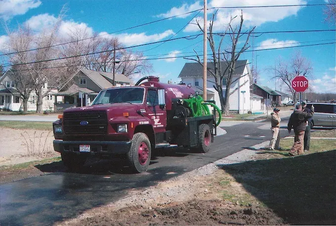 Maroon tow truck with green trailer on a suburban street near workers and a stop sign