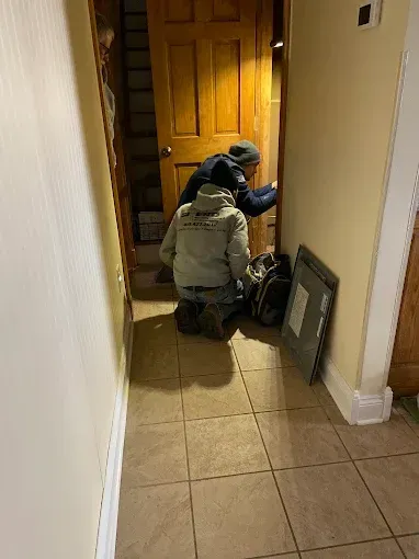 Person kneels by a front door in a hallway, reaching toward a bag next to a framed picture.