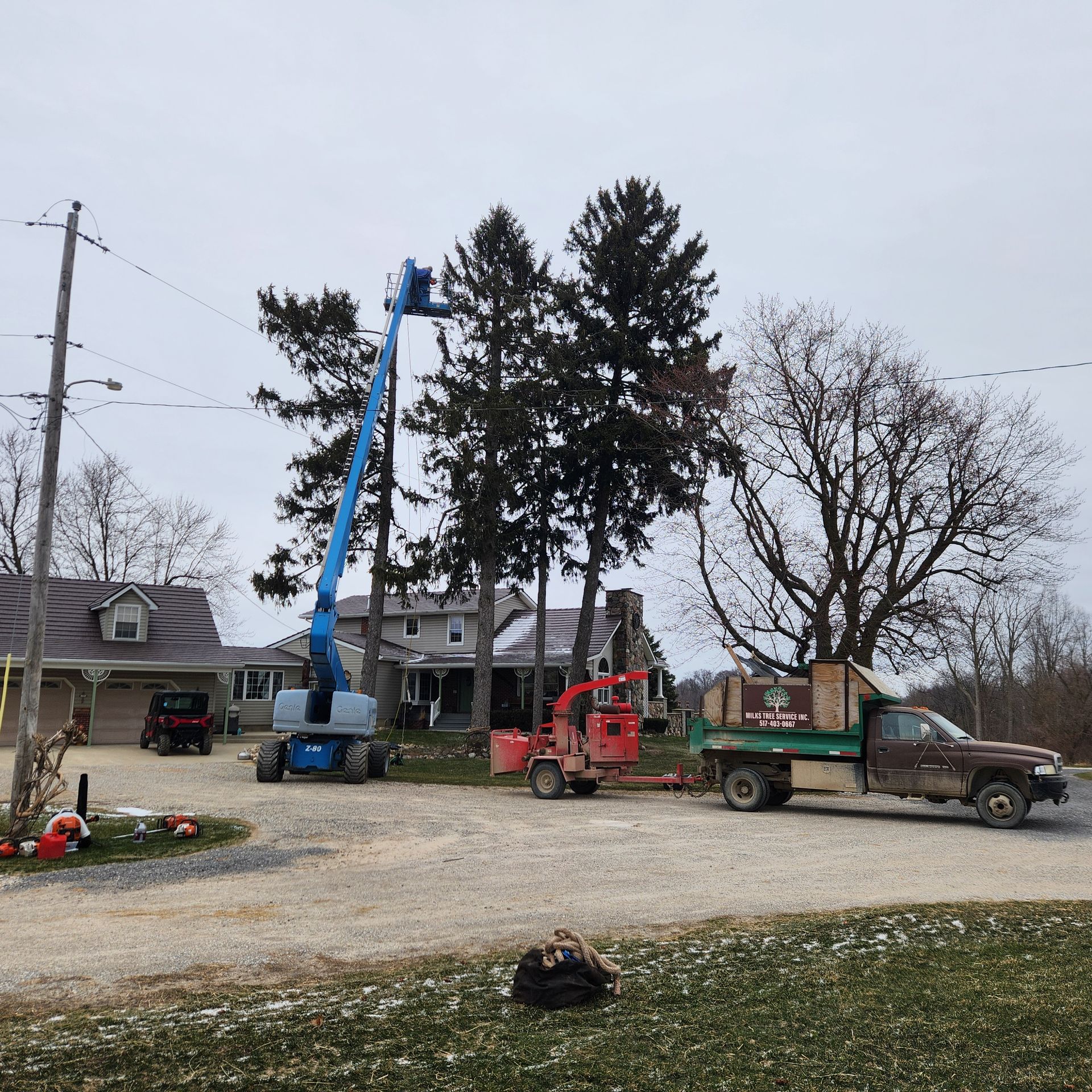 Utility trucks and a blue boom lift working beside a house and tall trees on a gravel lot