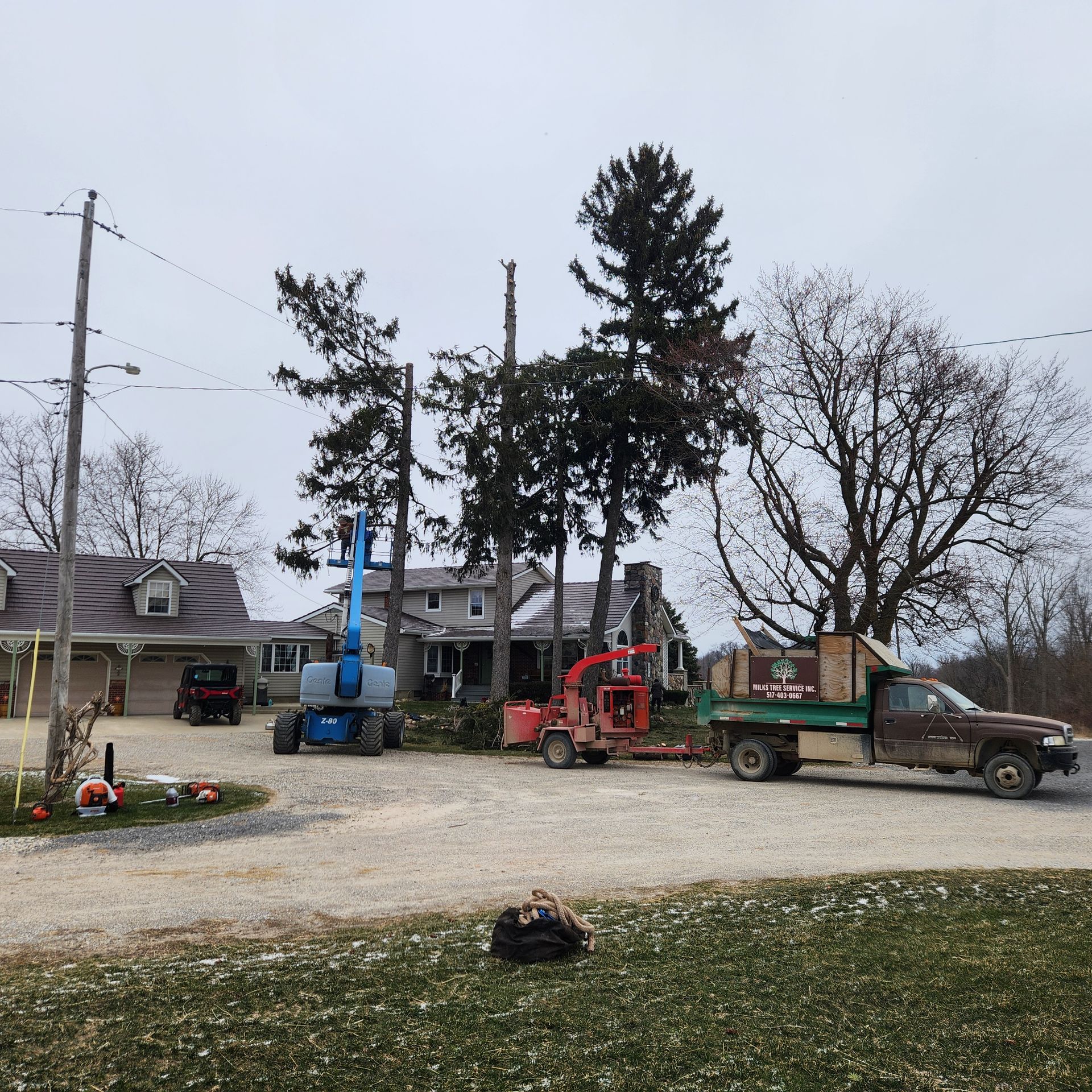 Landscape of a yard with trucks, a red lift, and tall trees beside a house on a cloudy day