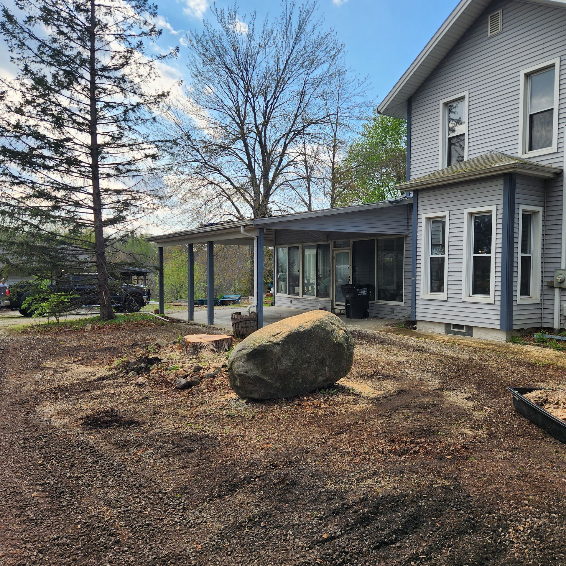 Side yard with a large boulder, covered patio, and gray house under a blue sky