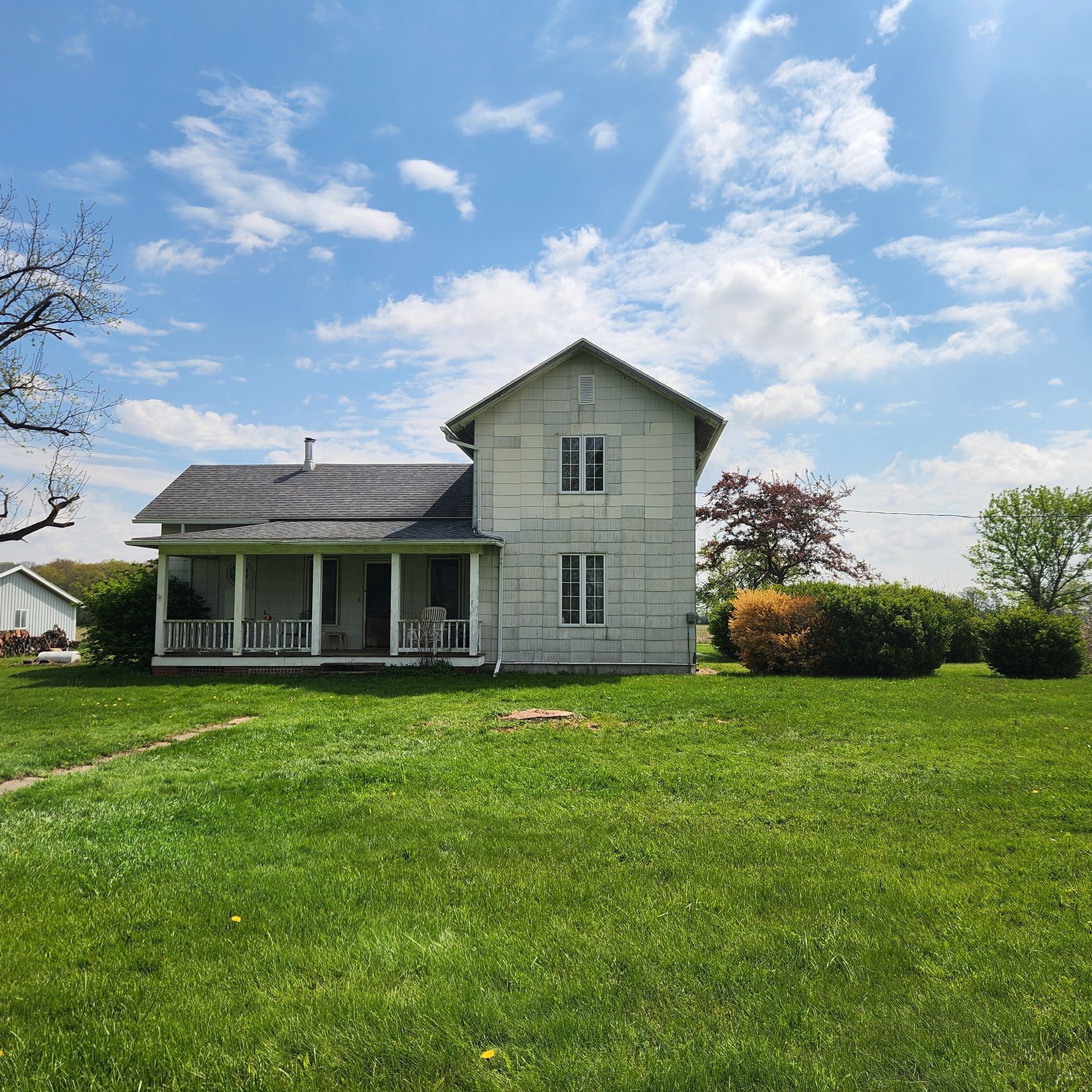 White farmhouse with porch beside a large green lawn under a bright blue sky with clouds
