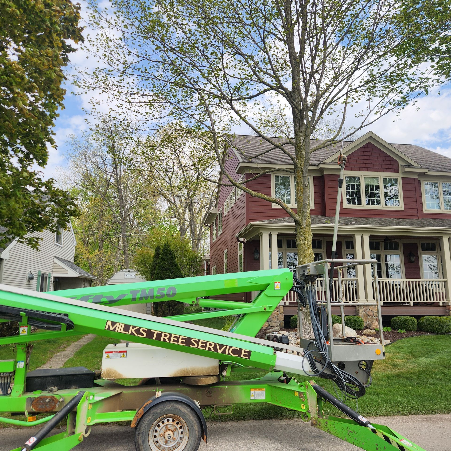 Bright green tree-service lift trailer in front of a red two-story house with a porch and trees.
