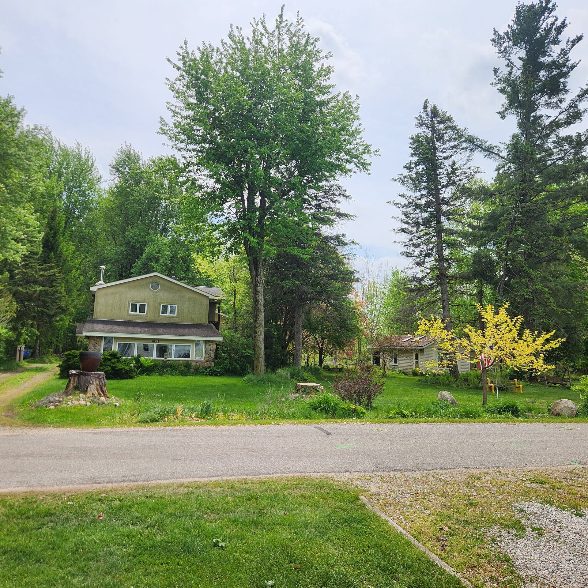 Suburban house with green lawn, tall trees, and a gravel driveway under a cloudy sky