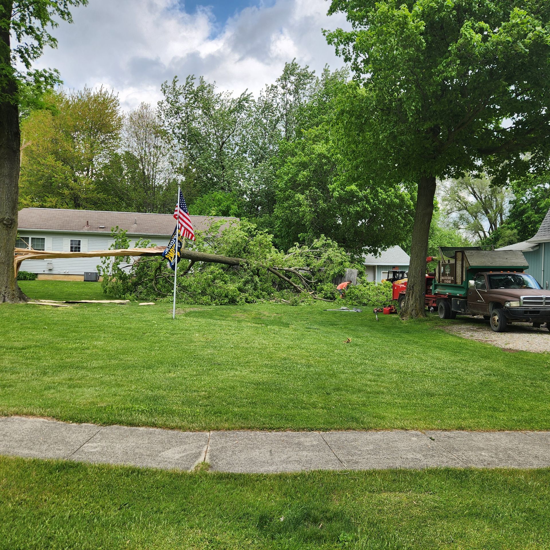 Suburban front yard with green lawn, American flag, trees, and a house with a carport and vehicle.