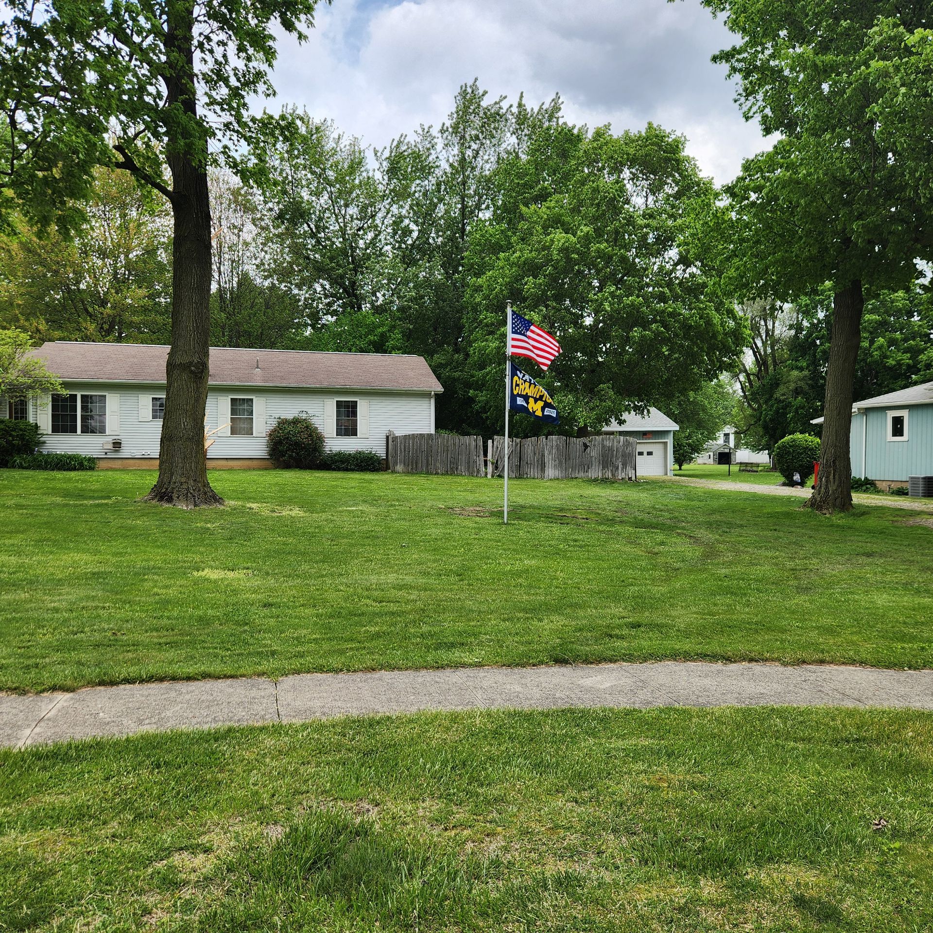 Suburban lawn with two flagpoles, American and yellow flags, trees, and single-story houses in the background