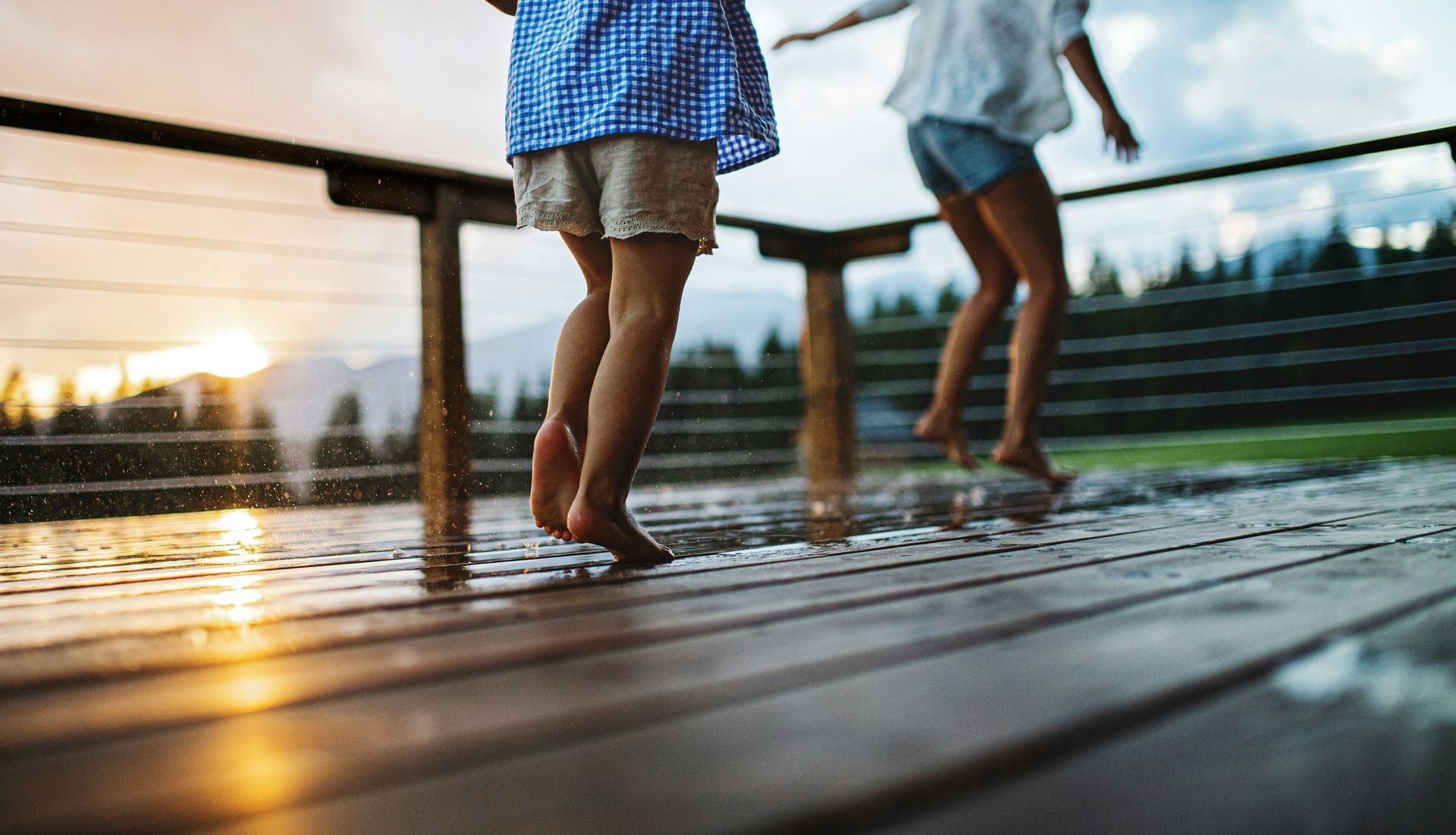 Two children in blue clothes walking barefoot on a wet wooden deck at sunset