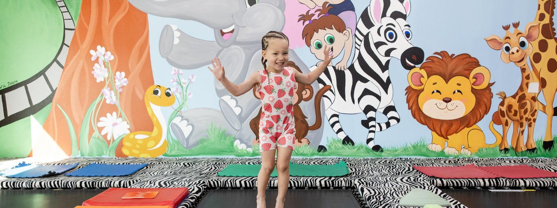 Young child in a leotard jumping on trampoline before a colorful jungle animal mural, including elephant, zebra, lion, and snake
