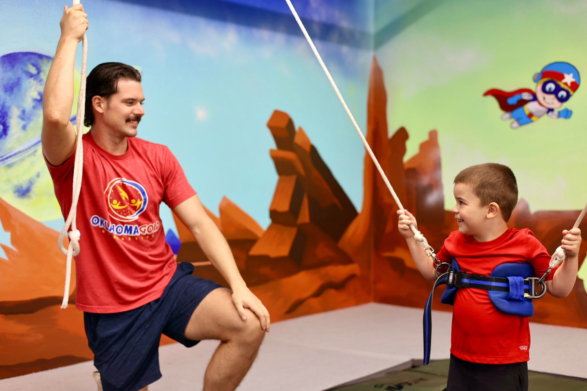 Adult and child on swing seats, smiling and facing each other in a colorful play area with a cartoon backdrop