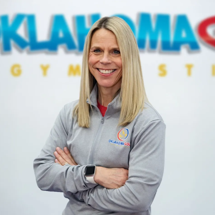Smiling woman with folded arms in gray Oklahoma Gymnastics jacket, standing before a logo backdrop