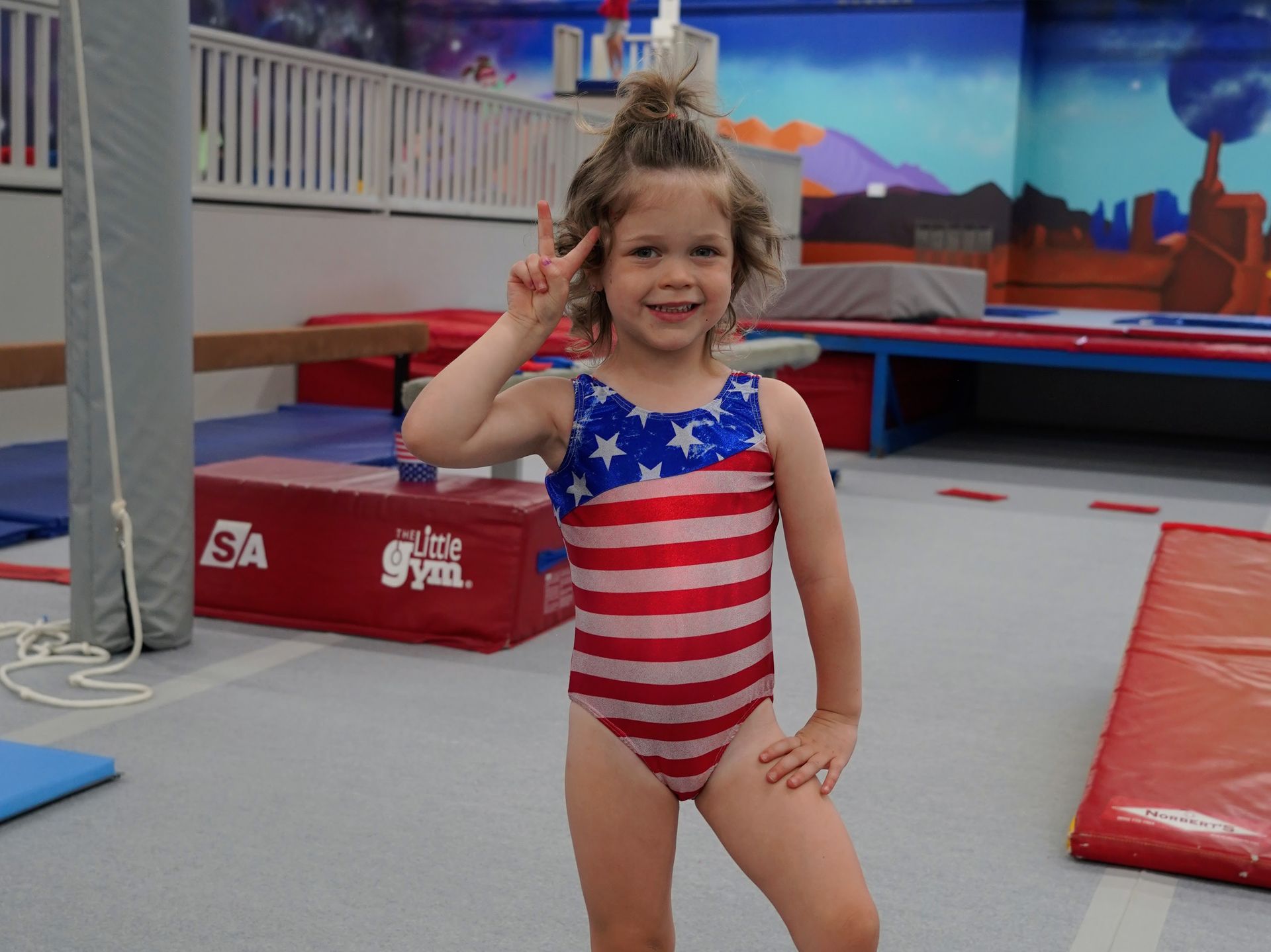 Child in a star-spangled leotard smiling on a gymnastics floor with one hand raised