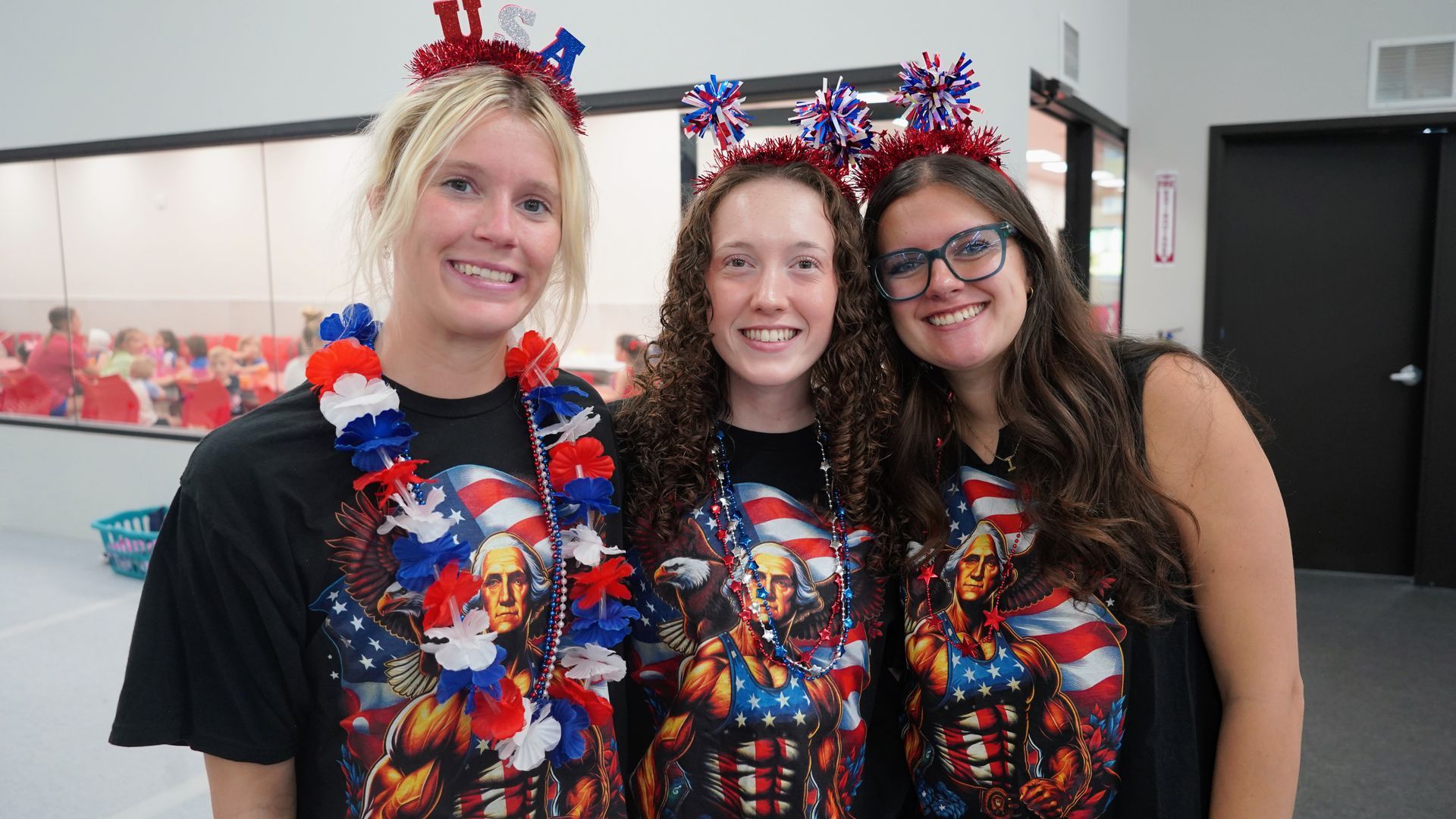 Three smiling people in matching red, white, and blue patriotic shirts and headbands stand indoors.