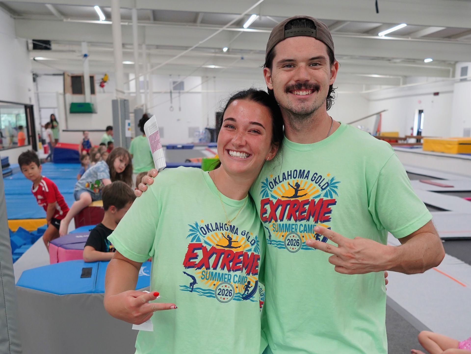 Two smiling people in matching green shirts pose in an indoor gym with children playing in the background