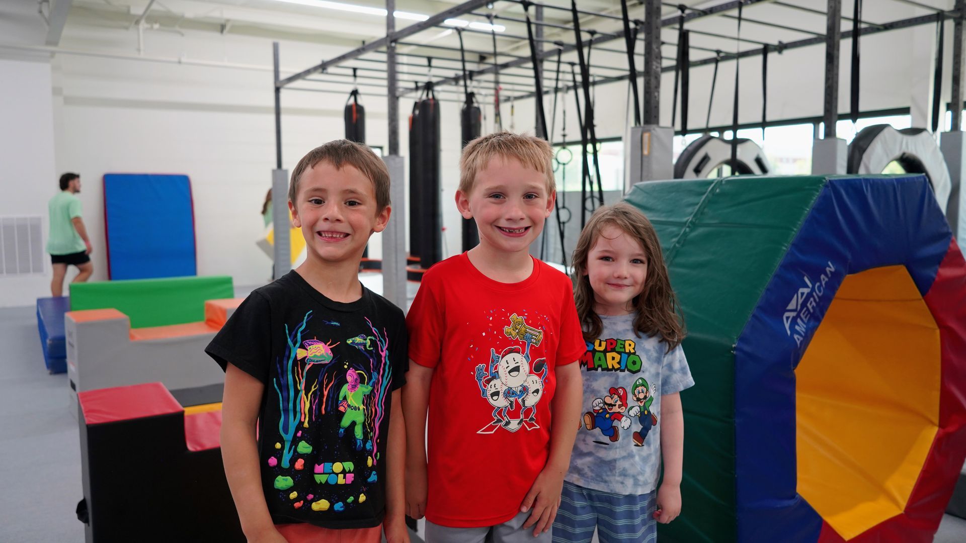 Three children smiling in a gym, standing by colorful padded equipment and mats.