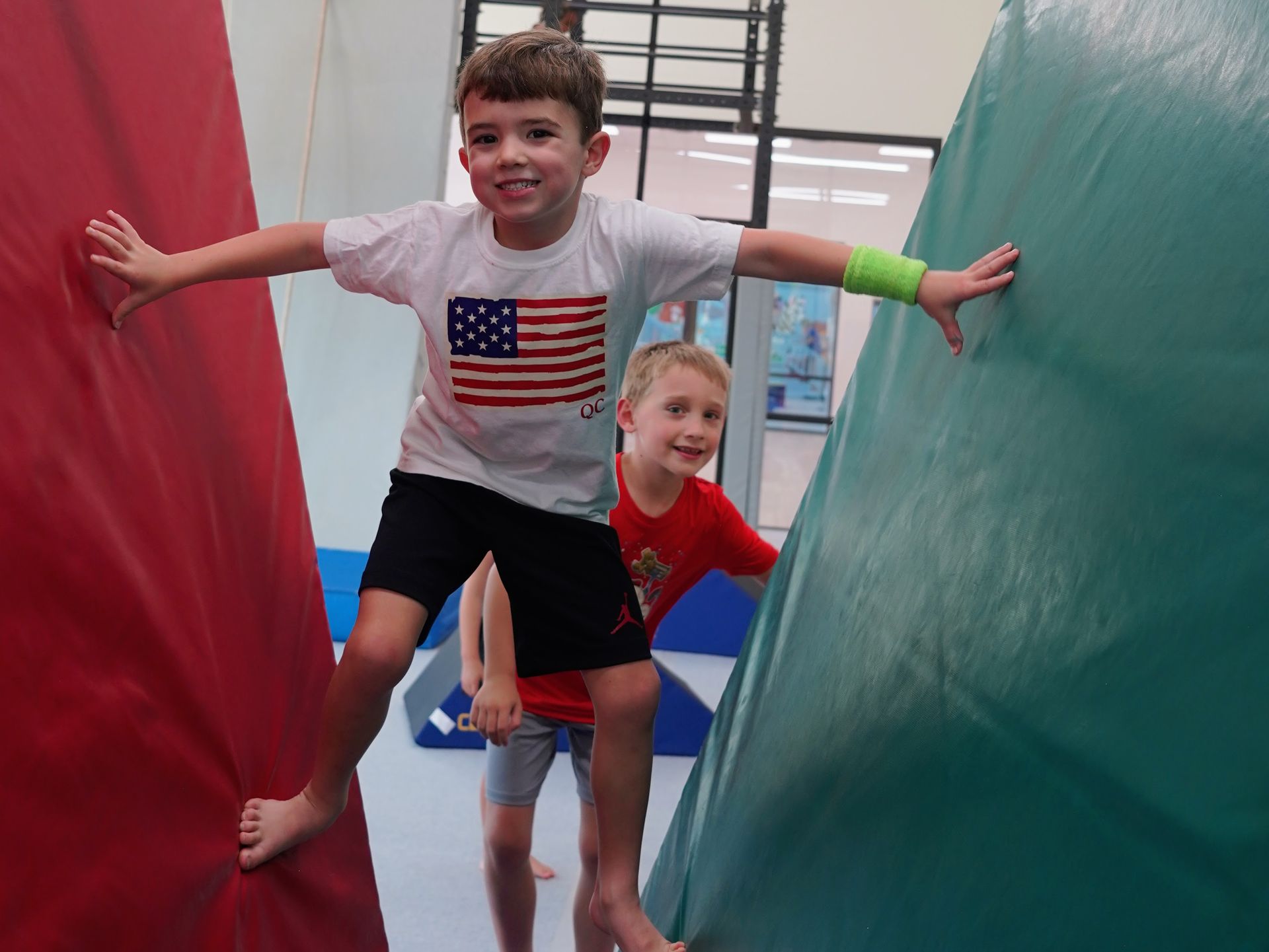 Two children smiling and balancing between red and green padded climbing walls indoors