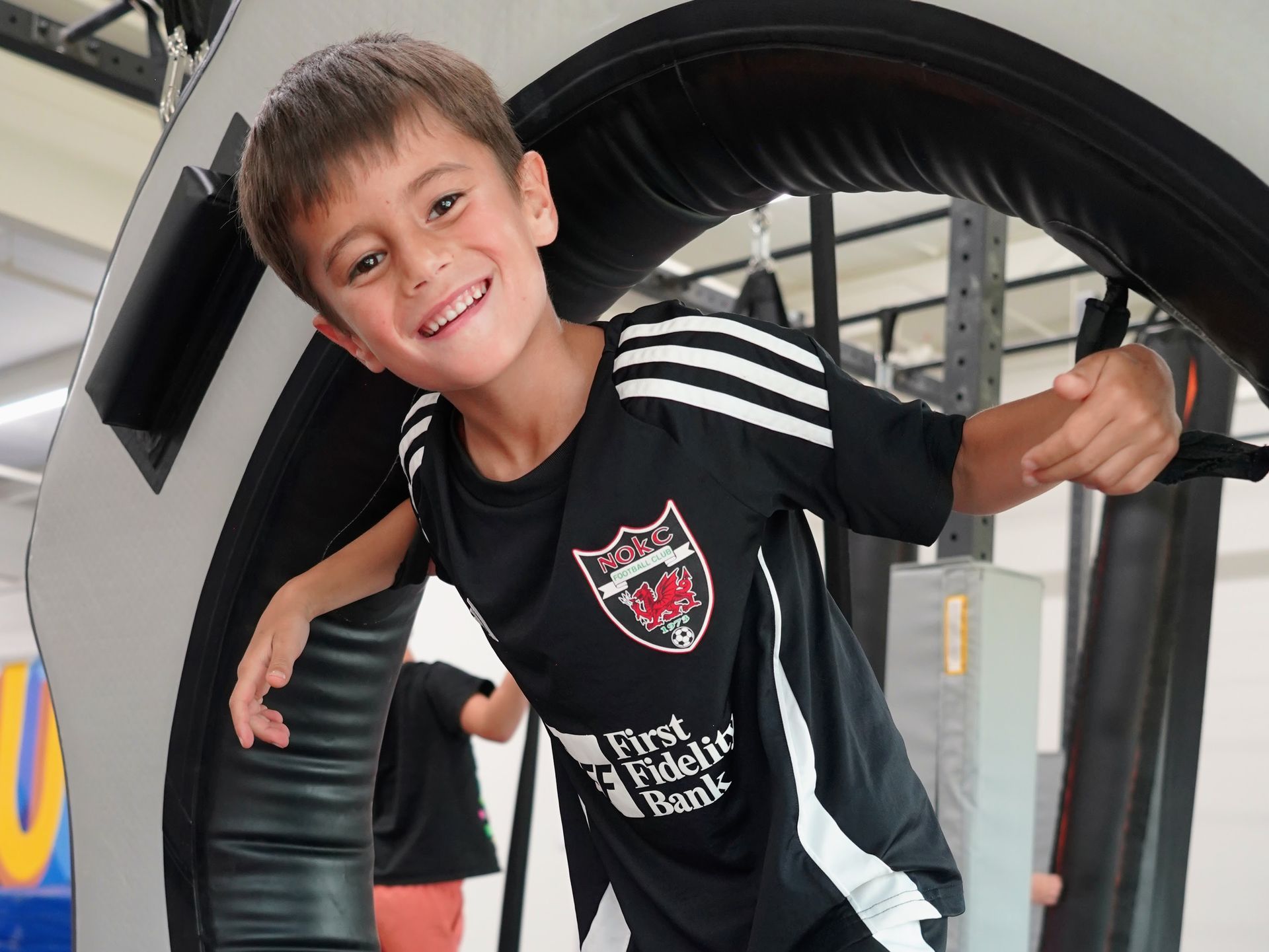 Smiling child in a black sports jersey leaning through a large tire obstacle indoors