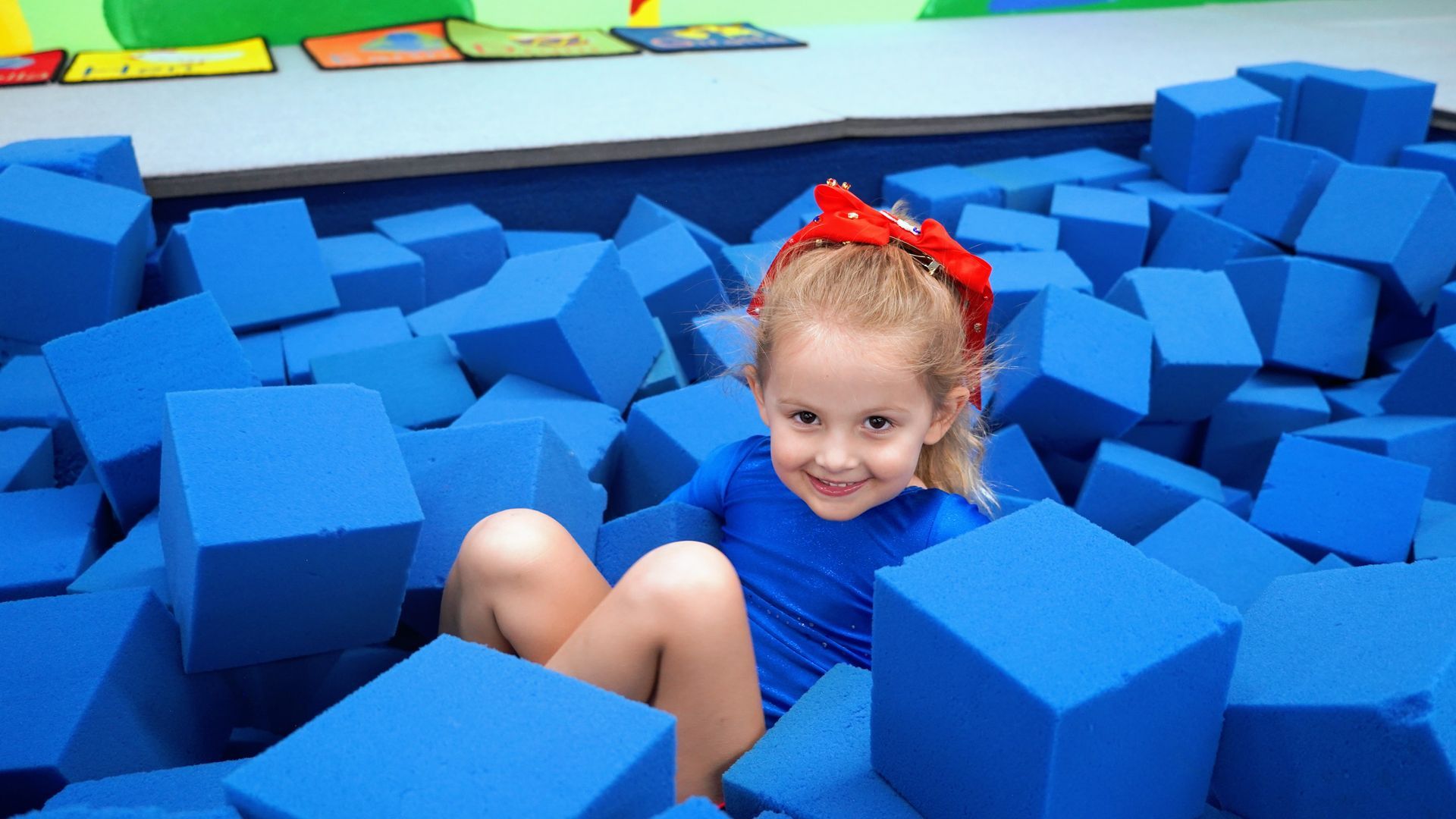Smiling child in a red bow lying in a blue foam pit surrounded by blue blocks